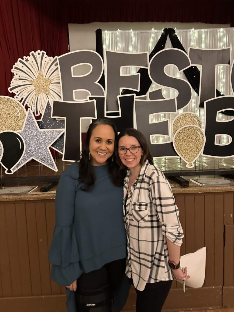 Two Bestowing the Basics volunteers smiling in front of an event backdrop
