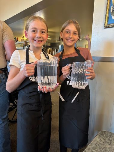 Two young volunteers in Bestowing the Basics aprons holding water pitchers at a community meal